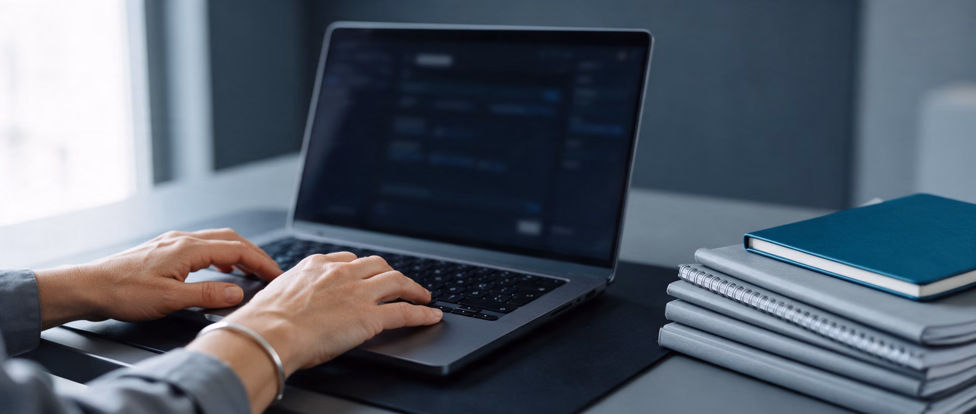 Close-up of a woman's hands on a laptop keyboard, the screen dark, beside a stack of grey spiral-bound documents topped with a teal notebook.