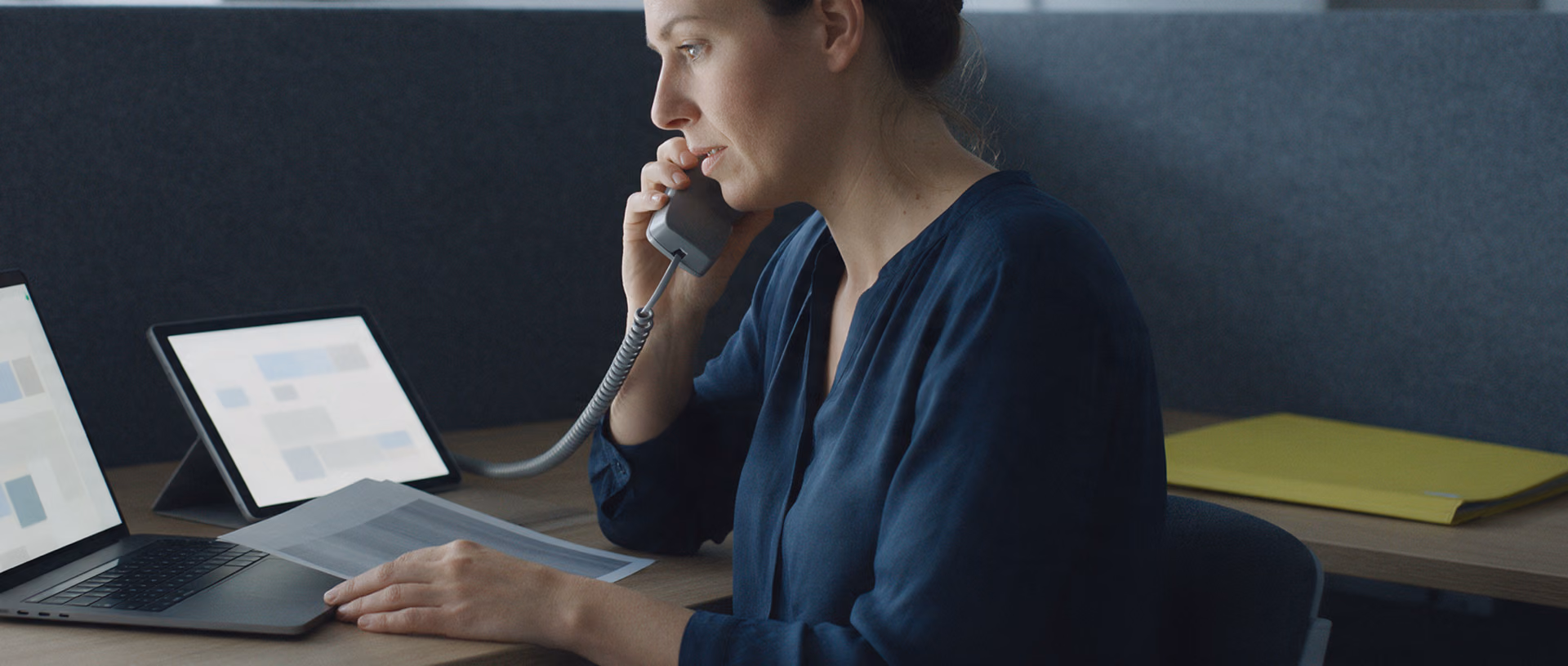 A woman at a back-office service desk holds a phone handset to her ear while looking from a laptop to a tablet, a printed sheet under her hand.