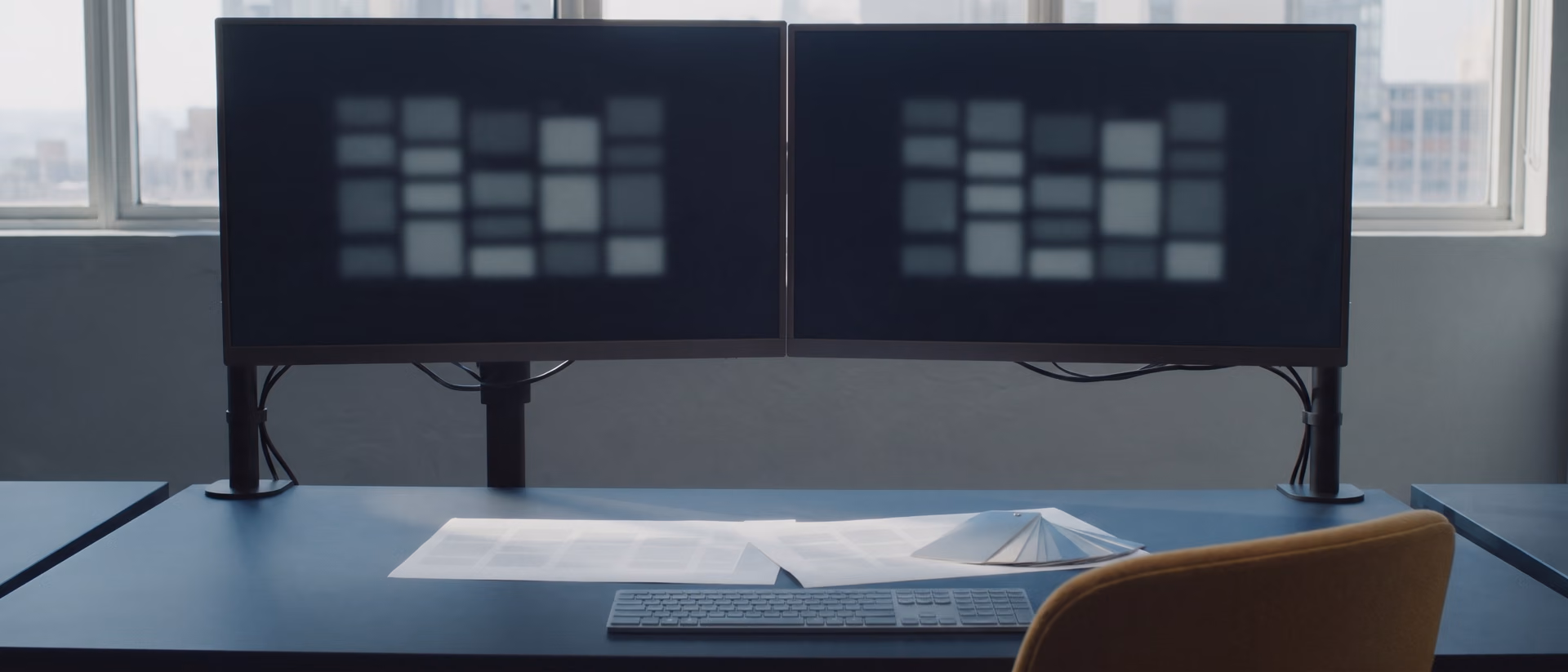 Two monitors on a dark desk showing the same blurred page grid, with printed layout sheets and colour swatches.