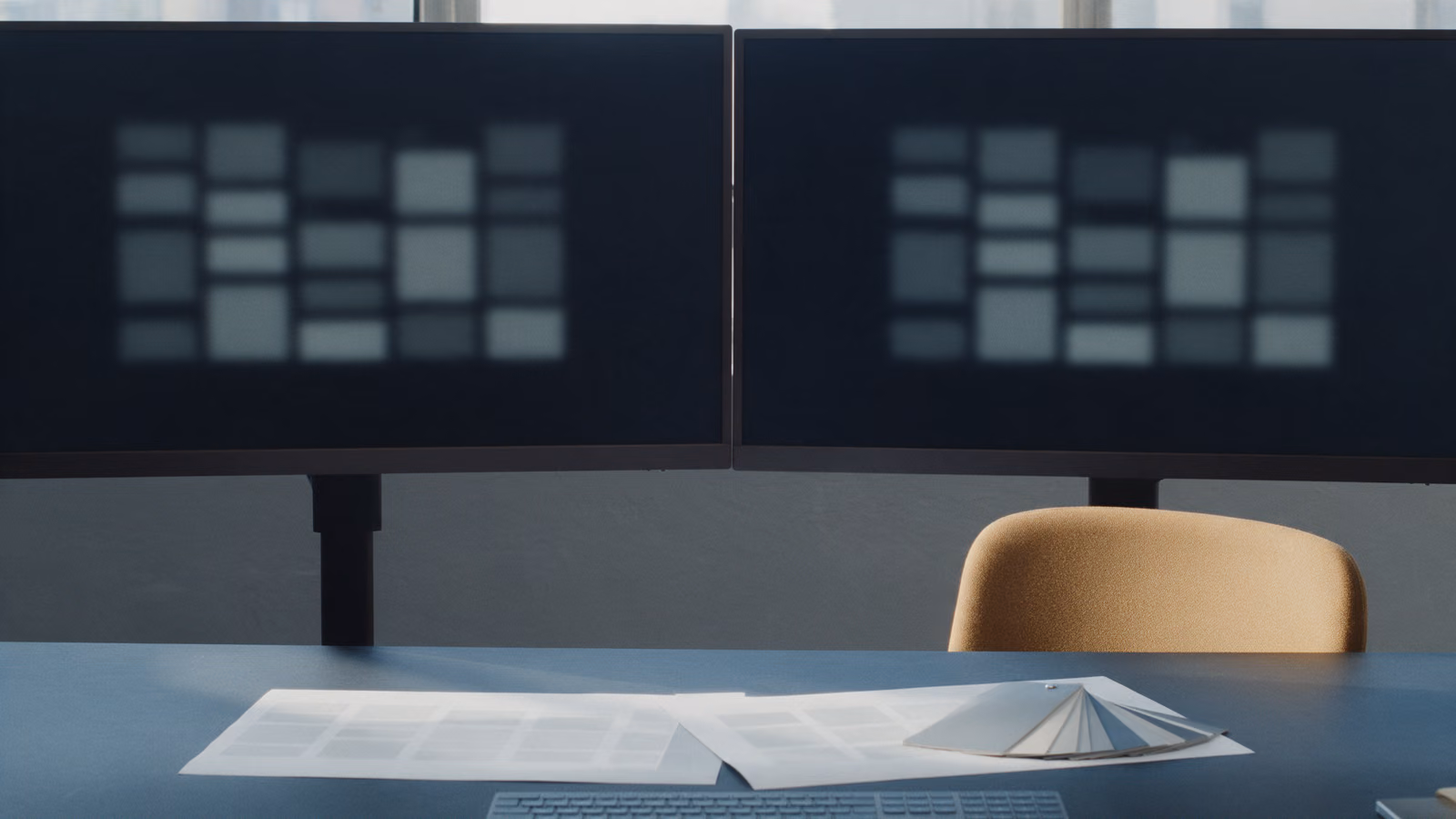 Two monitors on a dark desk showing the same blurred page grid, with printed layout sheets and colour swatches.