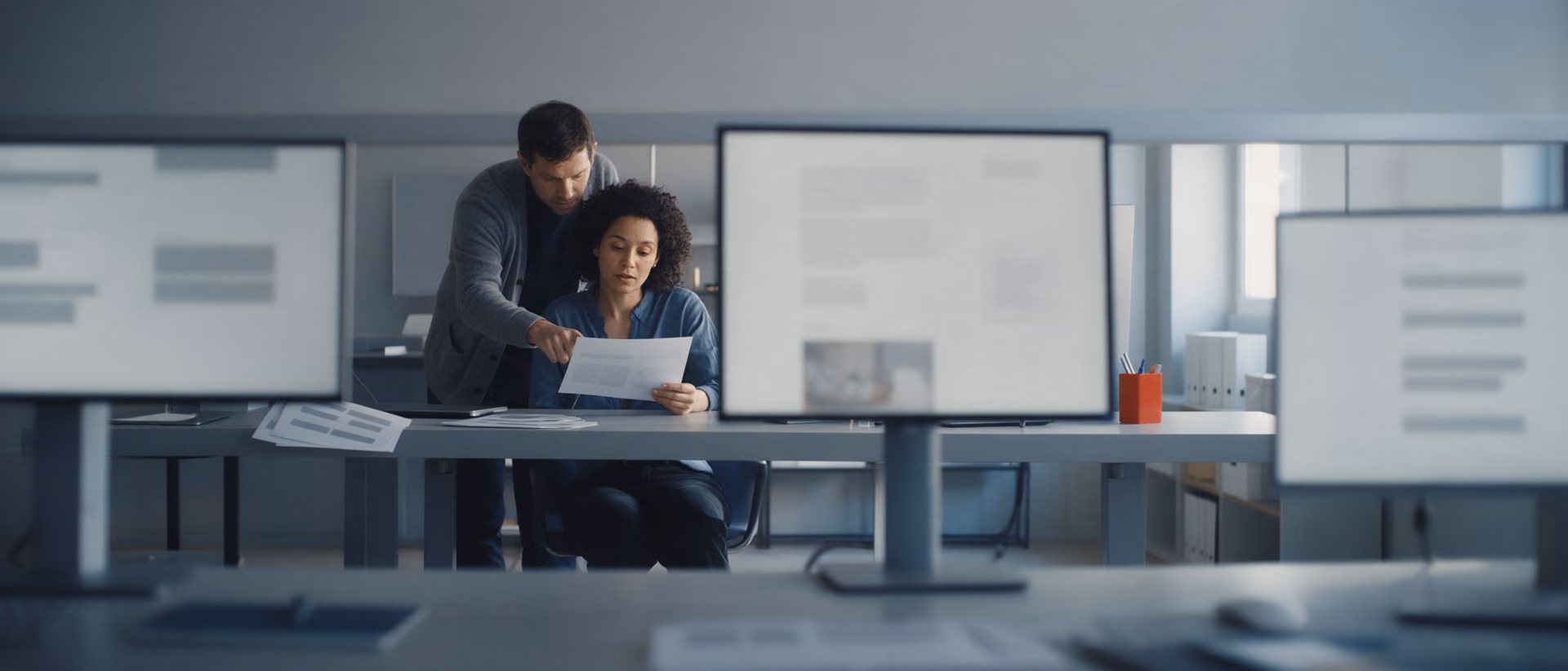 Two colleagues read a printed page beside a large monitor showing blurred paragraphs in a quiet editorial office, an orange pen cup on the desk.