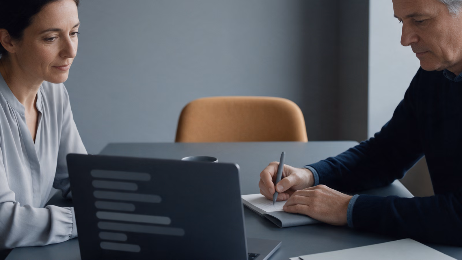 Two colleagues at a meeting table with a laptop, one writing notes.
