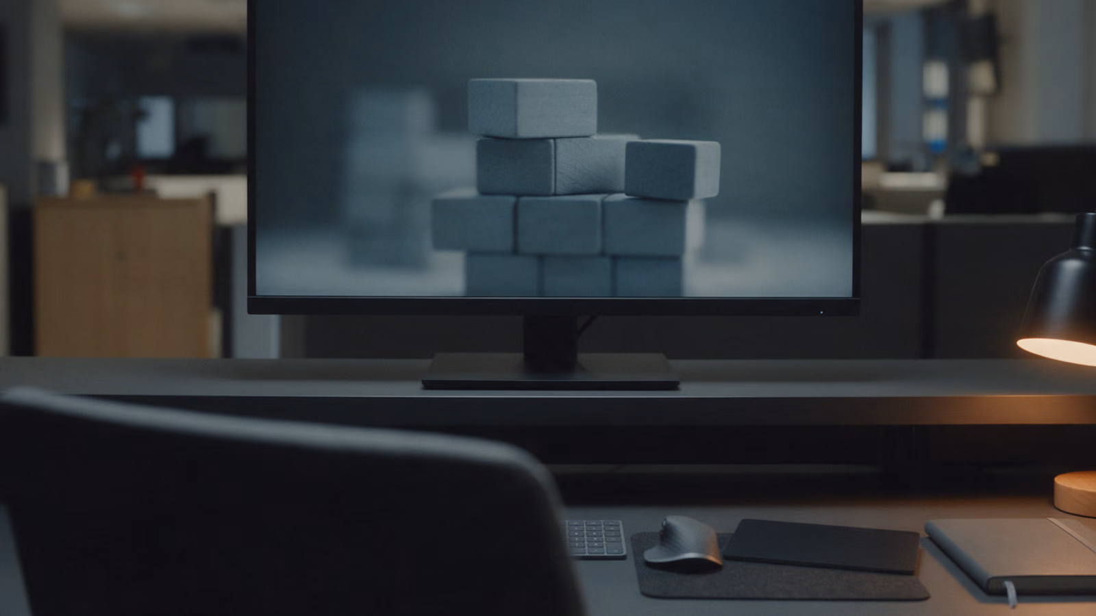 Monitor on an empty desk at night beside a lit desk lamp, its dark screen showing blurred stacked blocks.