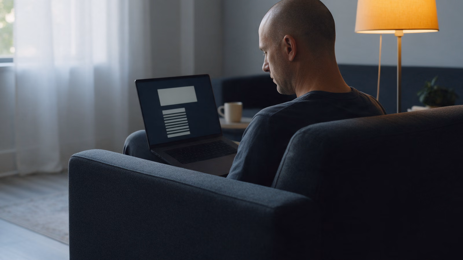 A man reading alone on a laptop in an armchair in a quiet lounge, a floor lamp beside him.