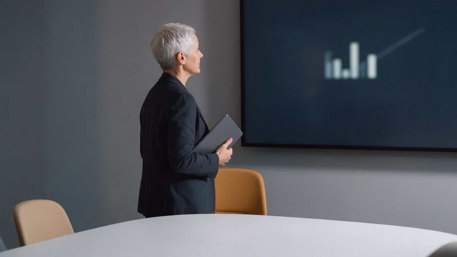 A woman with a tablet stands facing a wall screen showing blurred bars of unequal height in a quiet meeting room.