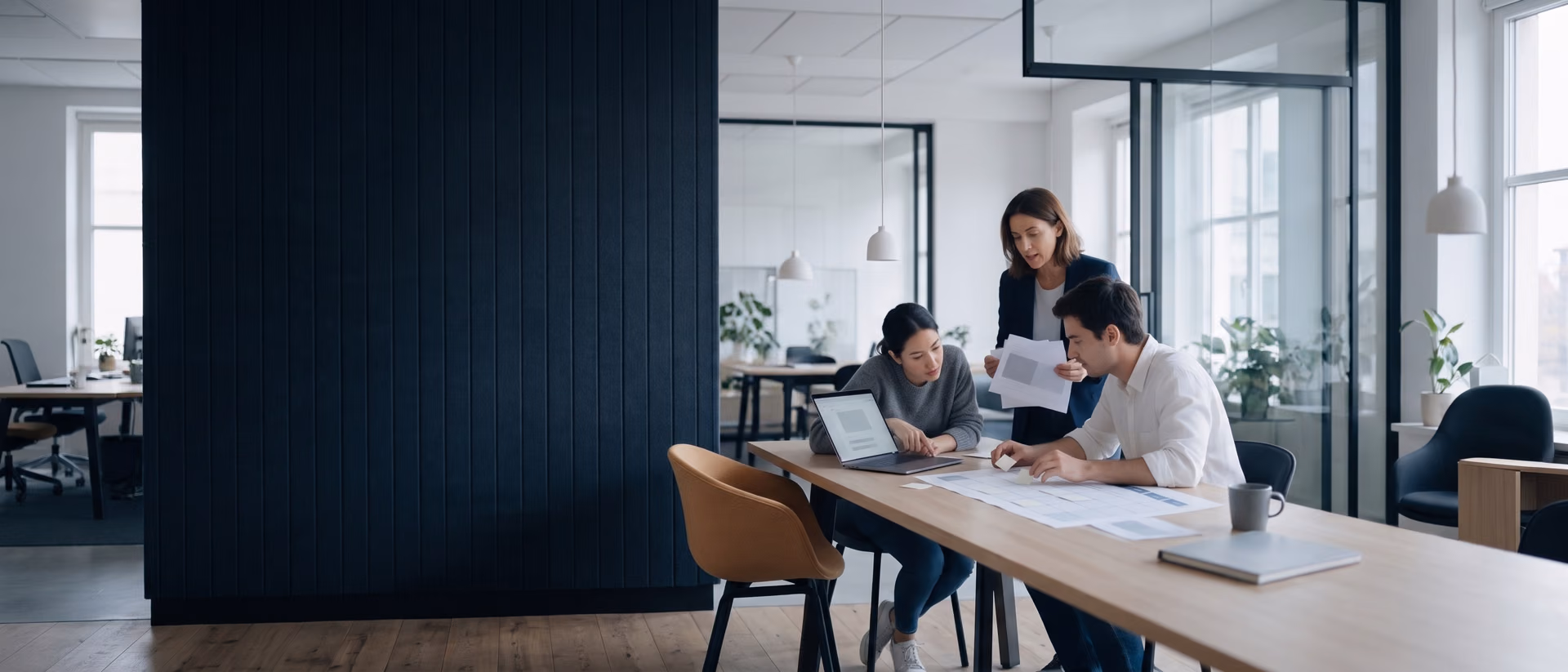 Three marketing colleagues plan a campaign at an oak table: one reviews an email draft on a laptop, one places sticky notes on a printed content calendar, and a third stands holding landing page drafts.