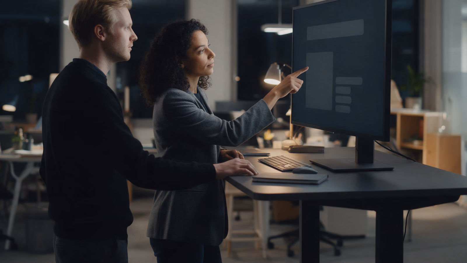 Two colleagues standing at a high desk after dark, one pointing at a dark monitor.