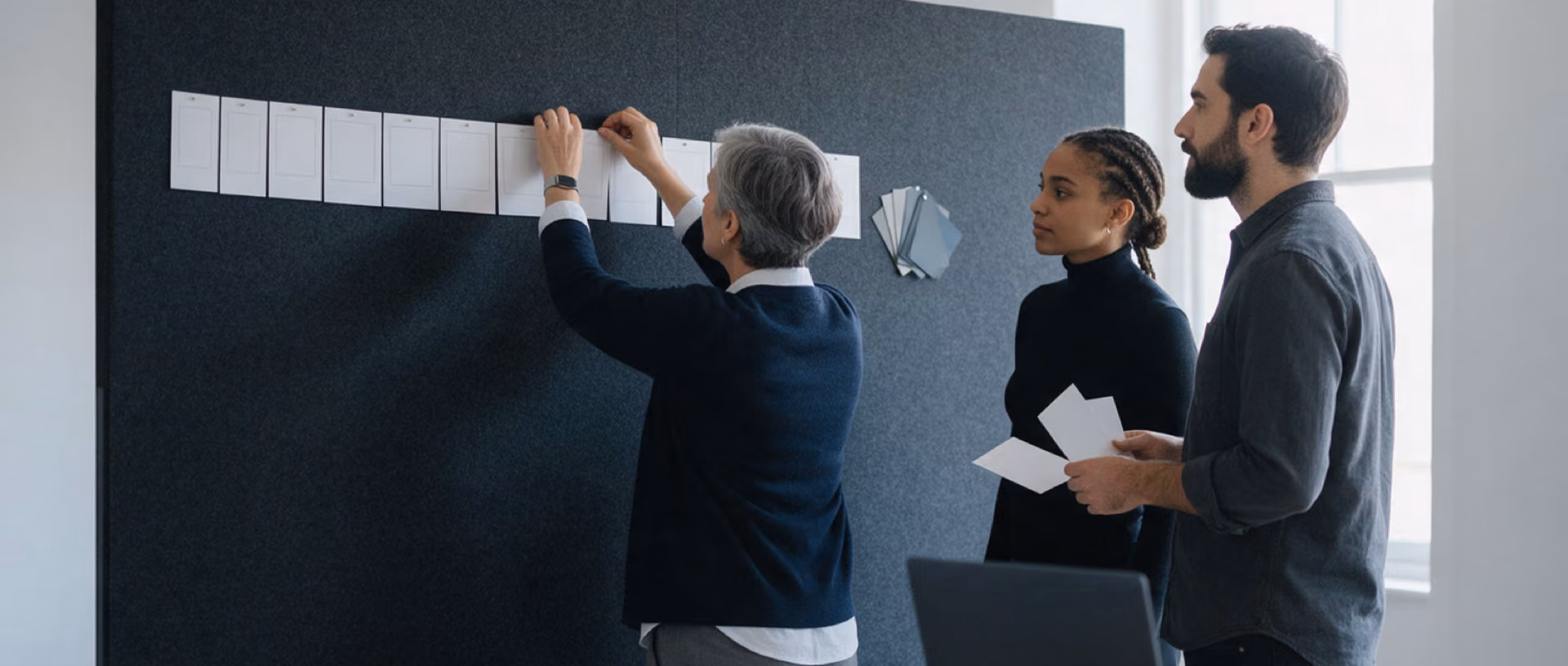 Three colleagues pinning blank layout cards to a dark wall in a project room.