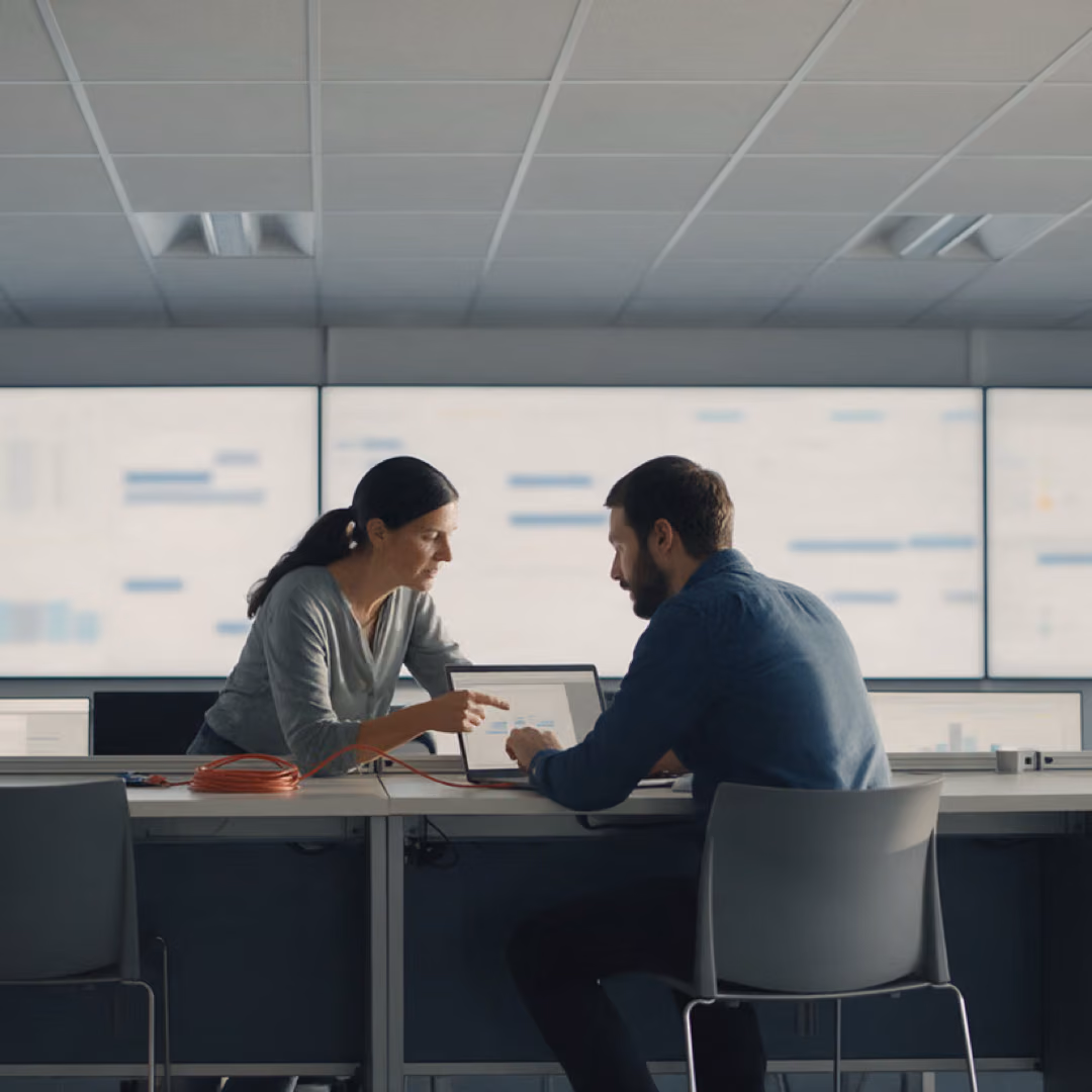 Two colleagues sit at a table with a laptop in front of a wall of large screens showing charts.