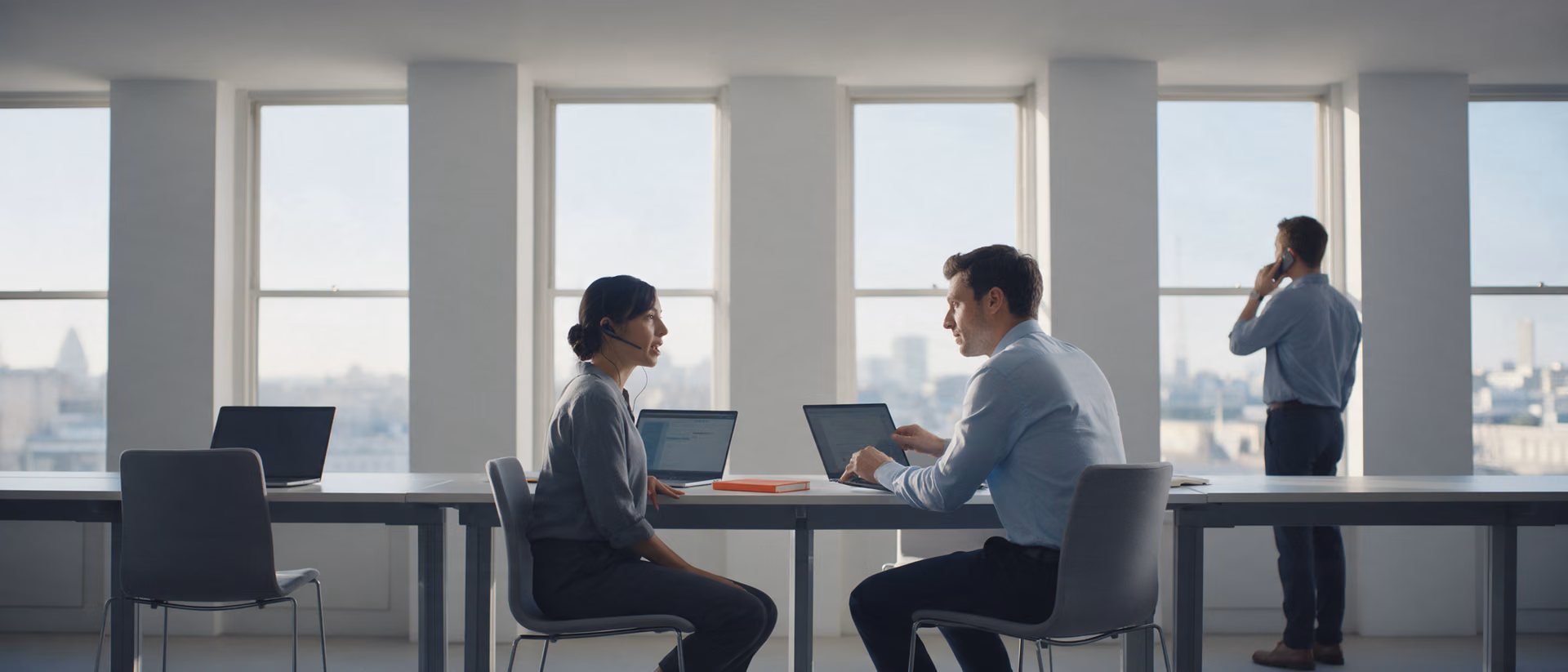 Two salespeople talk across a long shared desk on a bright sales floor, one wearing a headset, a colleague on the phone at the tall windows behind, an orange notebook on the desk.