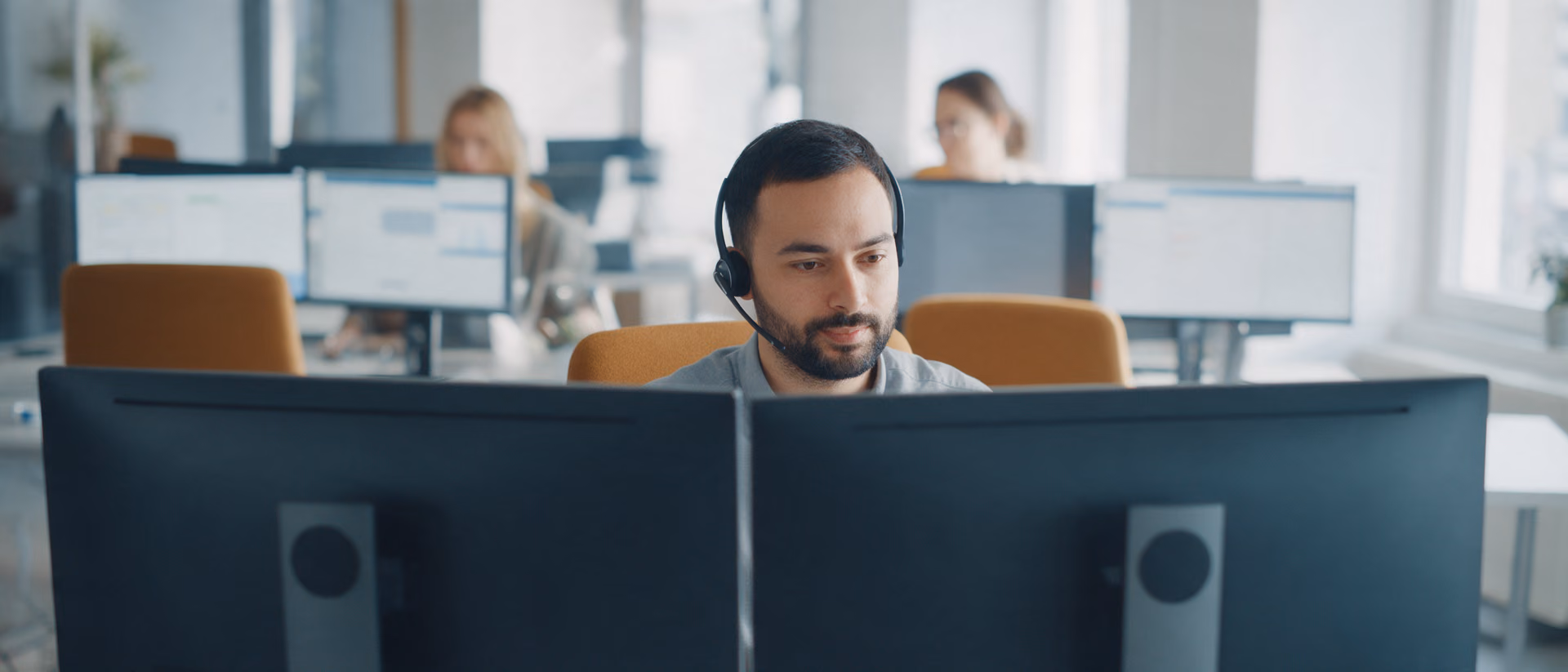 A service agent with a headset sits behind two monitors turned away from the camera, colleagues out of focus behind.