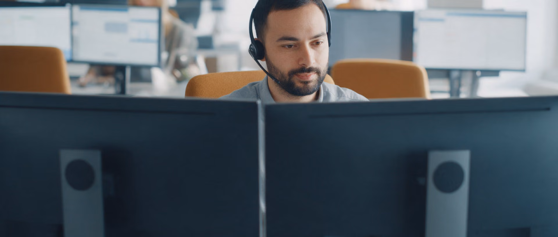 A service agent with a headset sits behind two monitors turned away from the camera, his hand on the mouse, colleagues out of focus behind him.