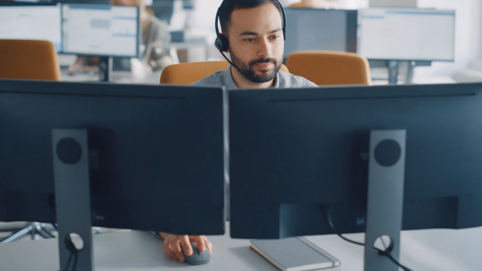 A service agent with a headset sits behind two monitors turned away from the camera, his hand on the mouse, colleagues out of focus behind him.