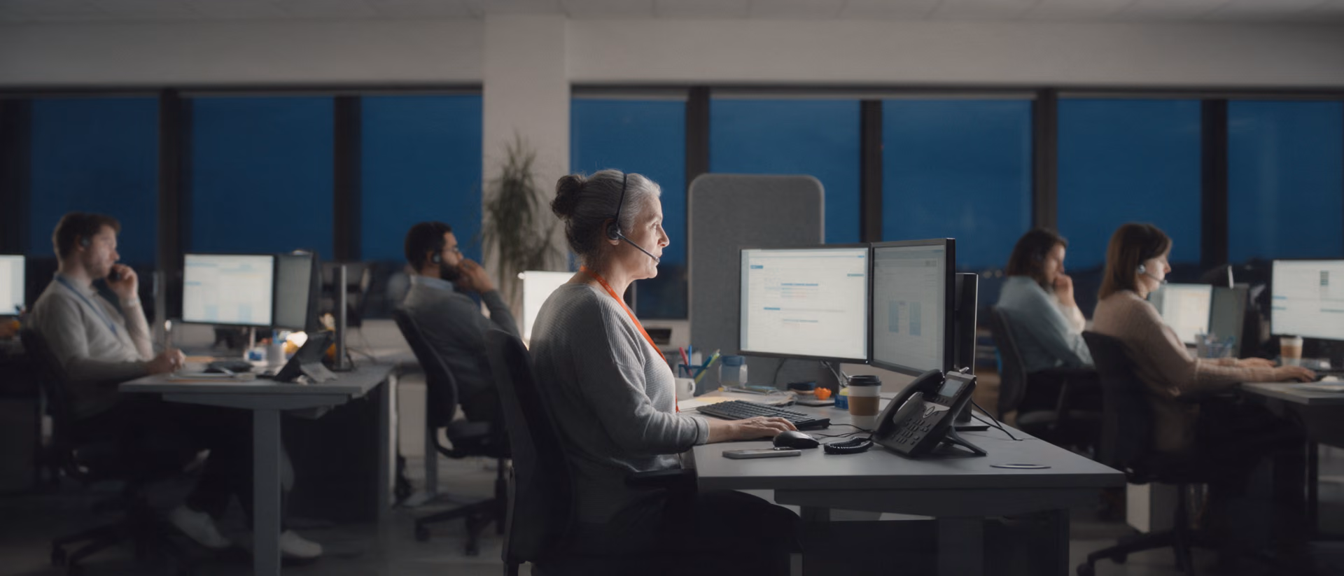 A service agent with a headset and an orange lanyard works at two monitors in a calm service centre after dark, colleagues at their desks on either side.