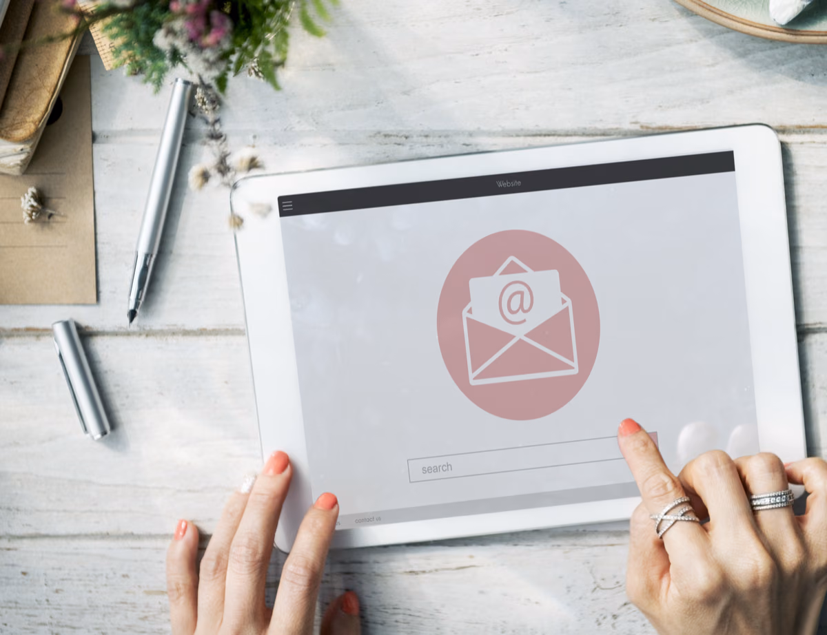 Zwei Hände halten ein Tablet mit einem E-Mail-Symbol über einem weißen Holztisch.