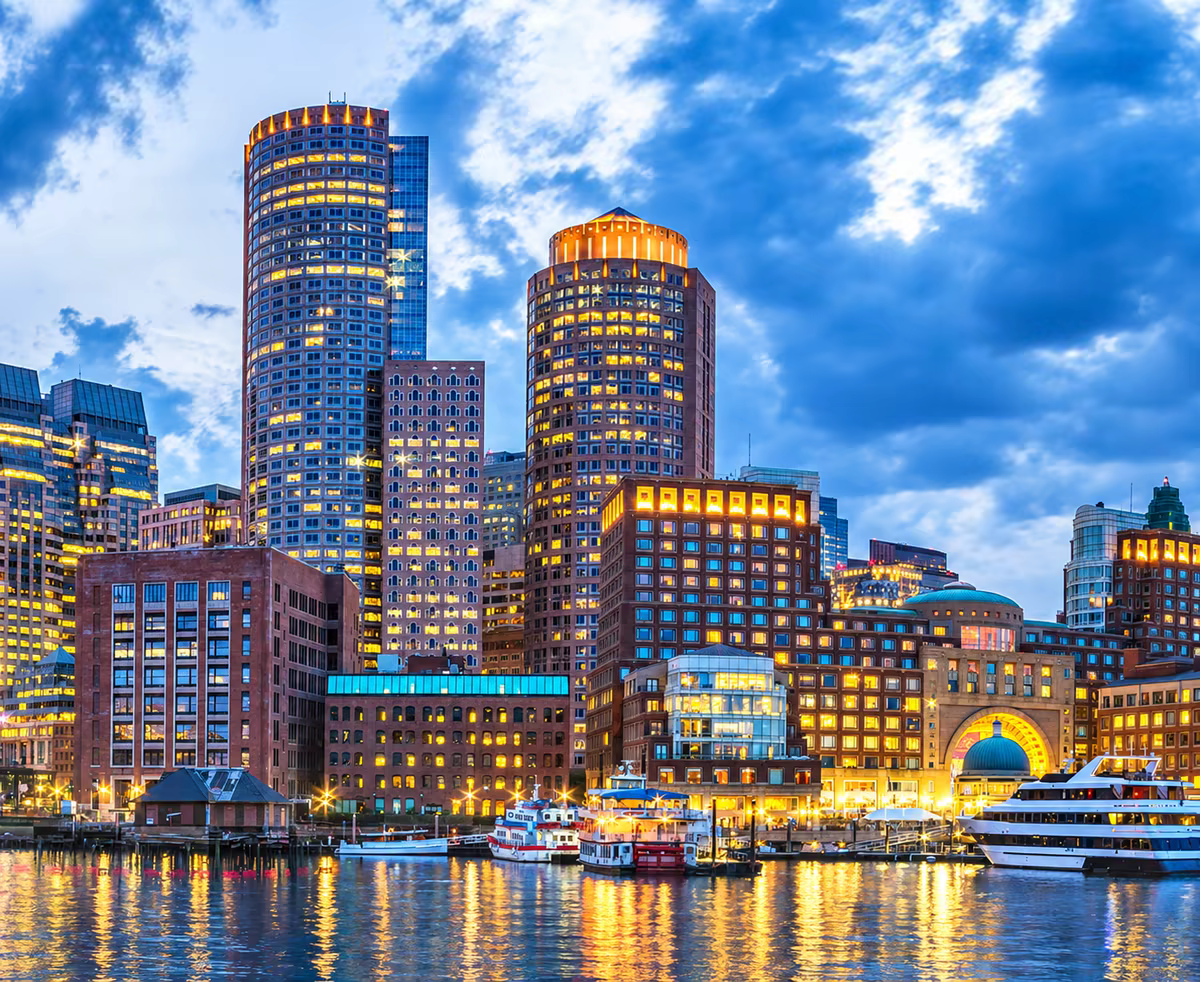 Die Skyline von Boston in der Dämmerung, über den Hafen gesehen, mit den erleuchteten Türmen des Finanzviertels, die sich im Wasser spiegeln.