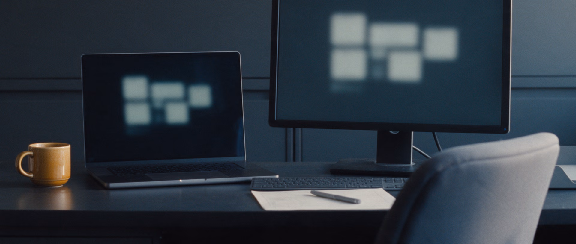 An unoccupied desk with an open laptop and a monitor side by side, both screens showing the same arrangement of blurred blocks, a mug at the corner and a letter tray at the right.