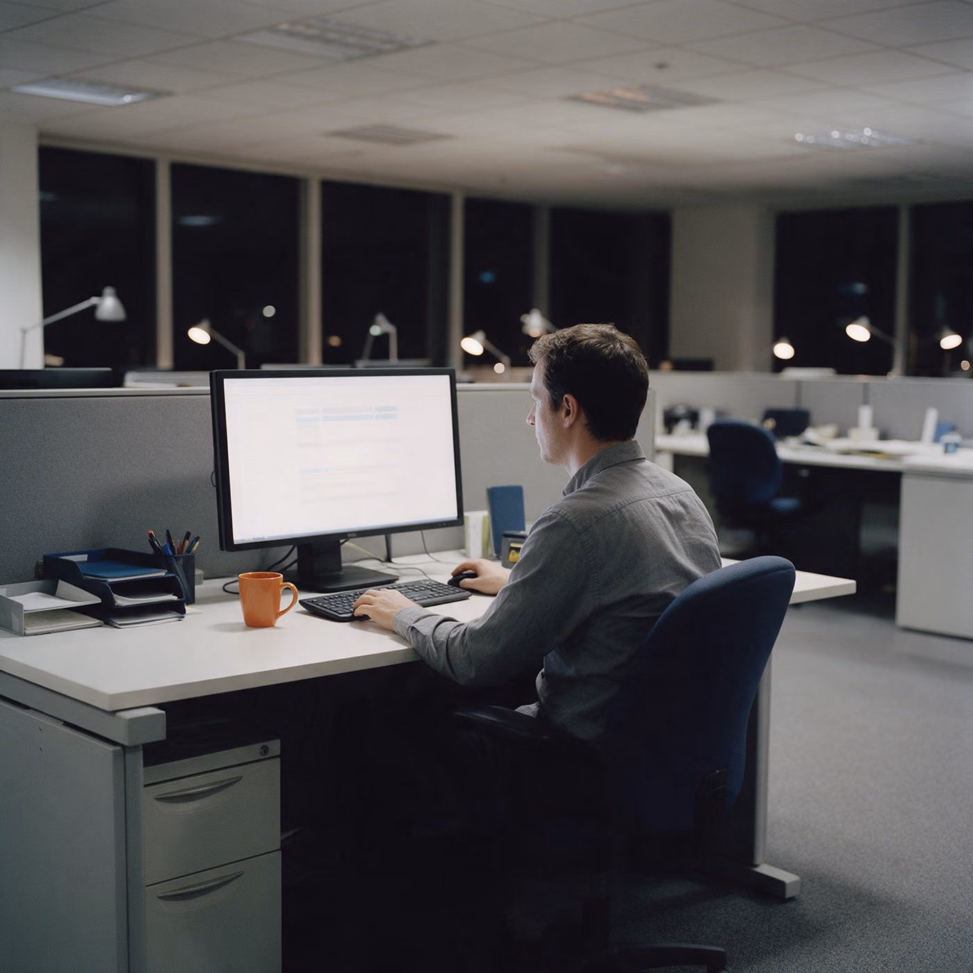 A man sits alone at a desk in an open office at night reading a single wide monitor, an orange mug beside the keyboard.
