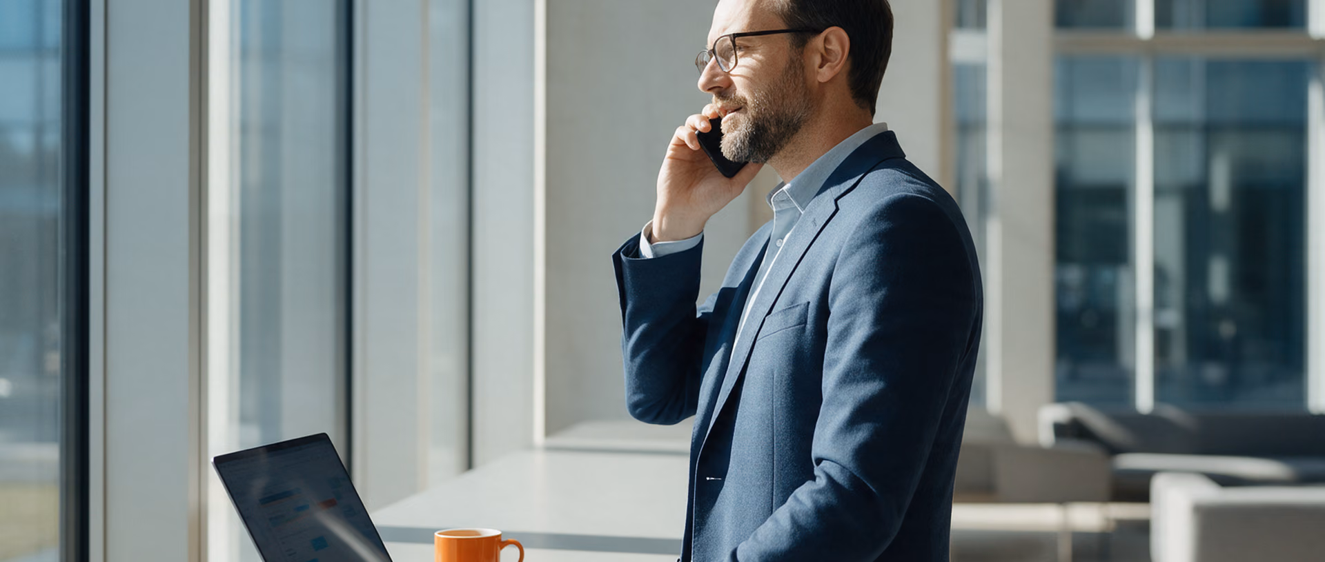A consultant stands at a high table in a bright client lobby, speaking on his phone while his laptop works on beside an orange mug.