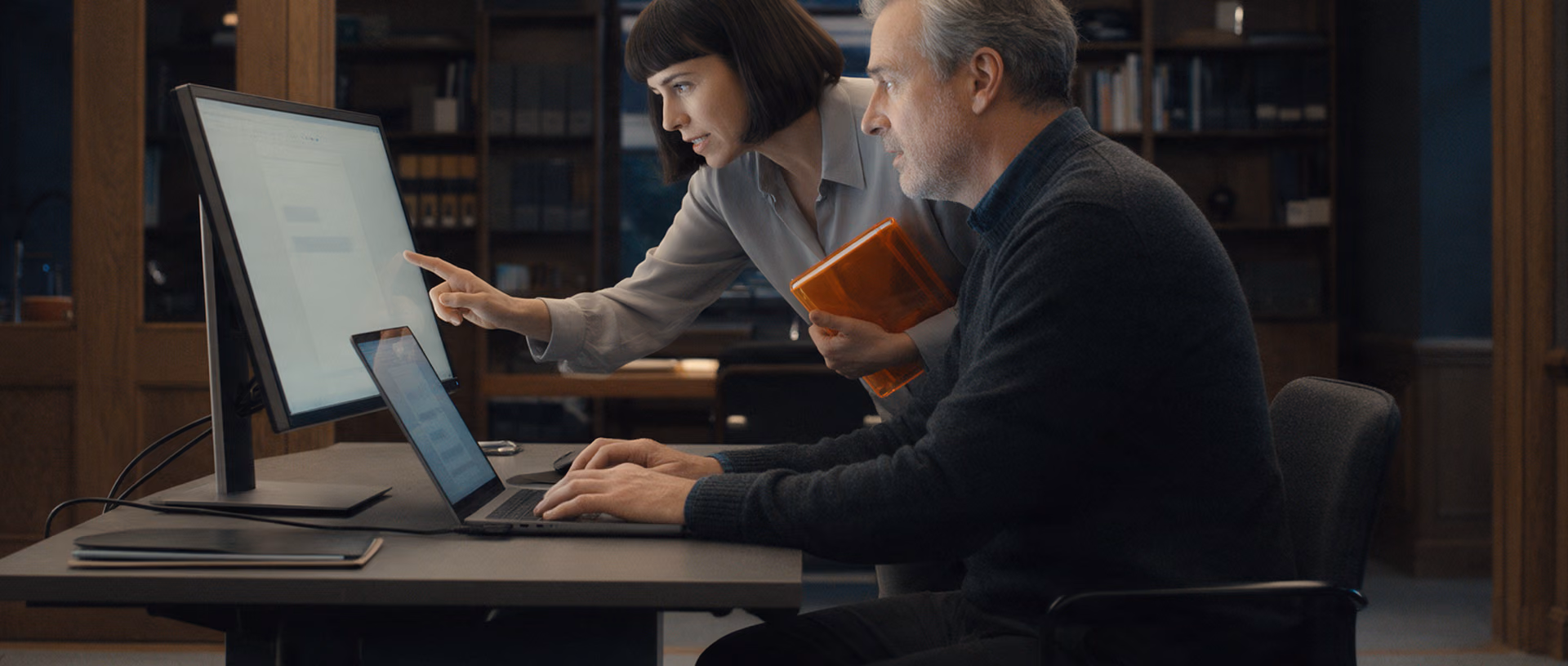A woman leans over a seated colleague's desk in a dark-panelled office after hours, pointing at his monitor while he types on his laptop.