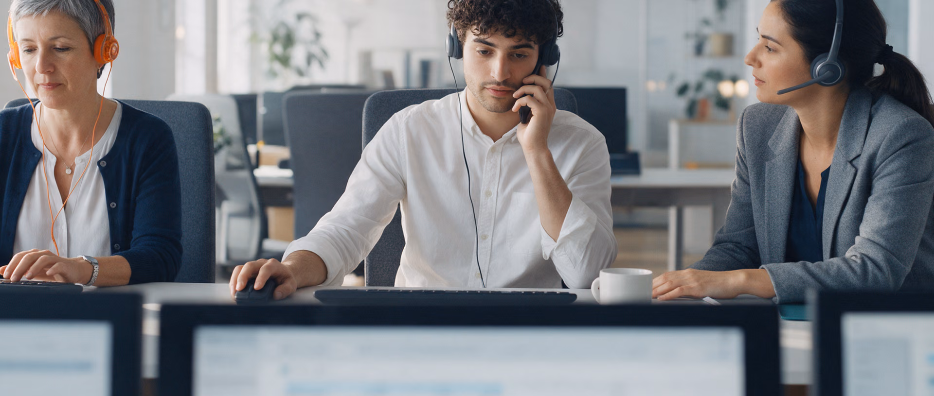 Three colleagues with headsets sit in a row at a customer service desk, one typing, one on the phone, the third turned towards him, three monitors in front of them.