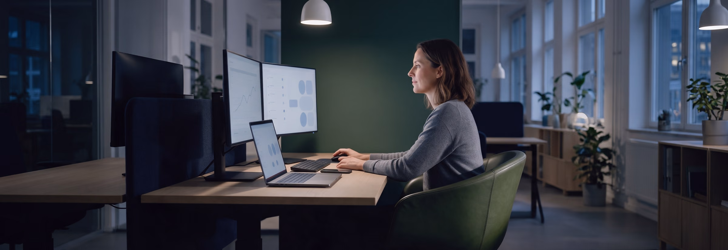An analyst in a green chair watches two monitors and a laptop with clean dashboards in the evening.