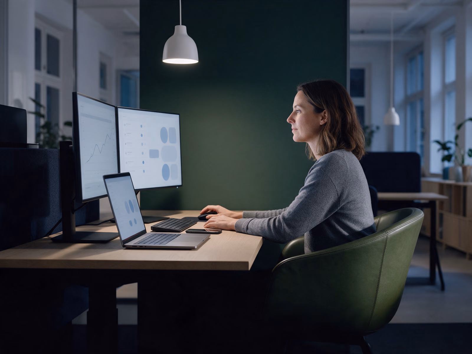 An analyst in a green chair watches two monitors and a laptop with clean dashboards in the evening.