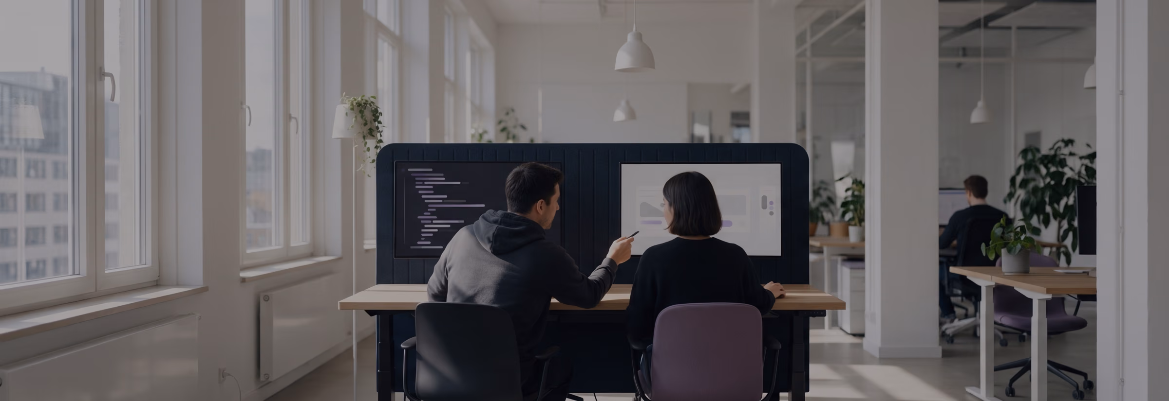 Two colleagues seen from behind at a shared desk, one pointing from his code editor at the design on her screen.