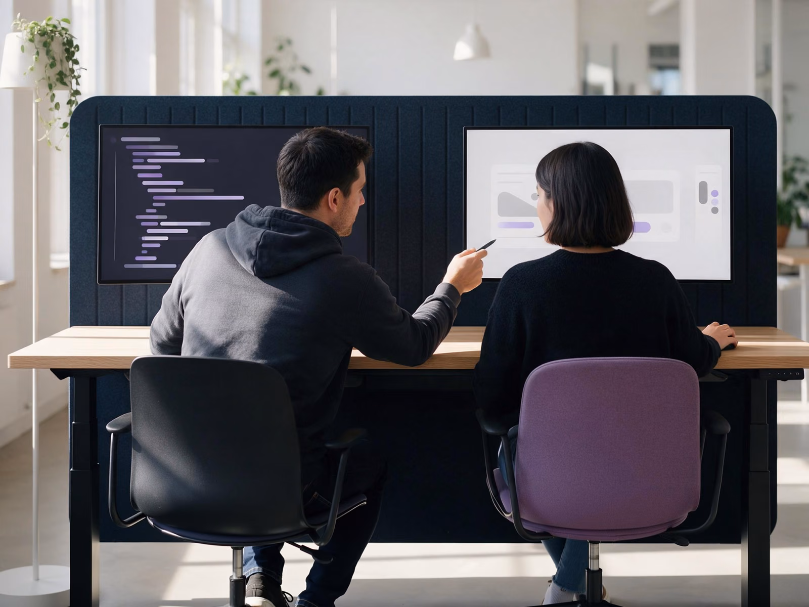 Two colleagues seen from behind at a shared desk, one pointing from his code editor at the design on her screen.