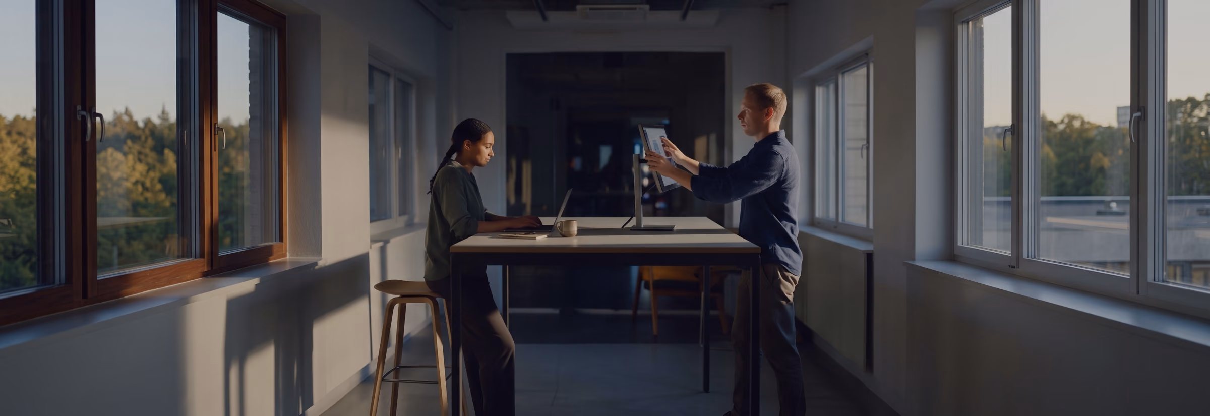 Two people at a standing desk, one typing on a laptop, the other turning a monitor on a stand towards her.
