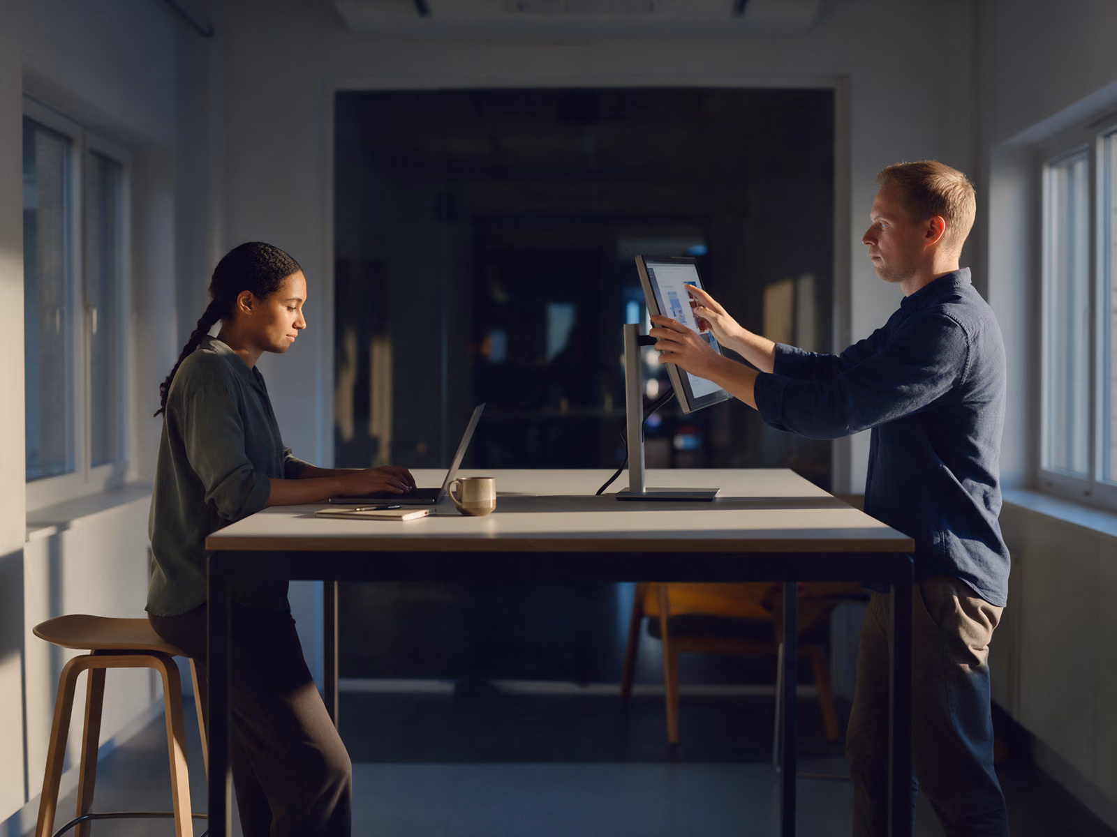 Two people work at one standing desk from opposite sides, one typing on a laptop, the other turning a portable monitor on a stand towards her.