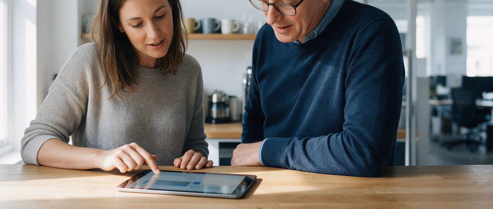 Two colleagues stand at a wooden counter in an office tea point, looking down at a tablet lying flat on the wood between them.