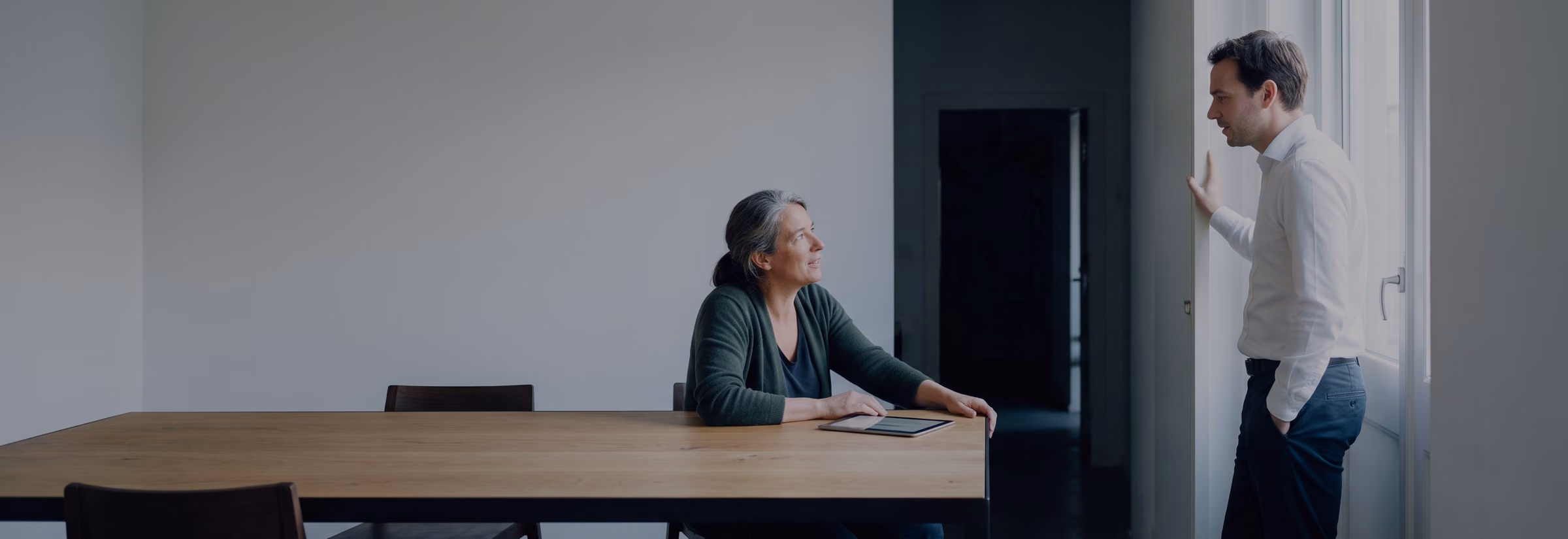 A woman with a tablet at a window-side table talking to a colleague standing by the window.