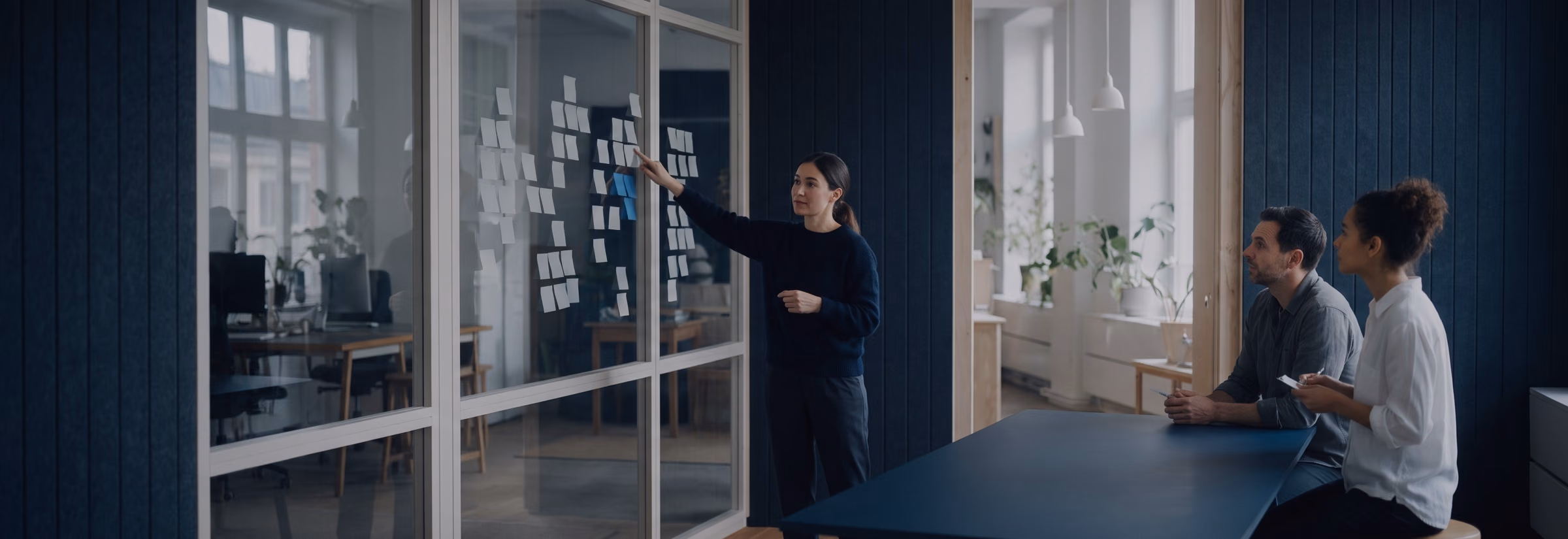 A workshop lead points at clusters of sticky notes on a glass wall while two colleagues follow.
