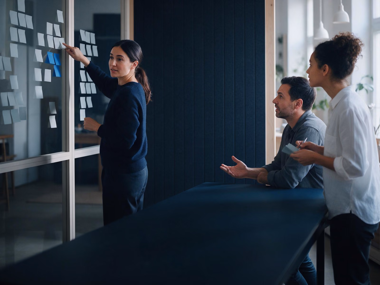 A workshop lead points at clusters of sticky notes on a glass wall while two colleagues follow.