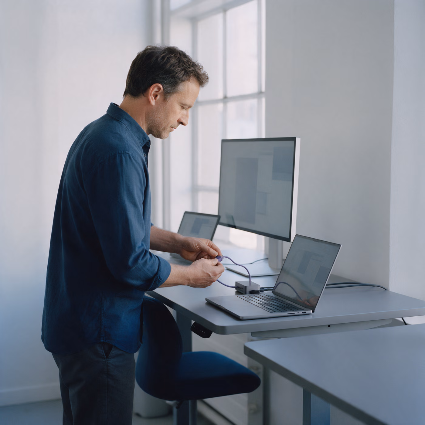 A man standing at a desk plugging a cable from a dock into a laptop, a monitor and a tablet beside it.
