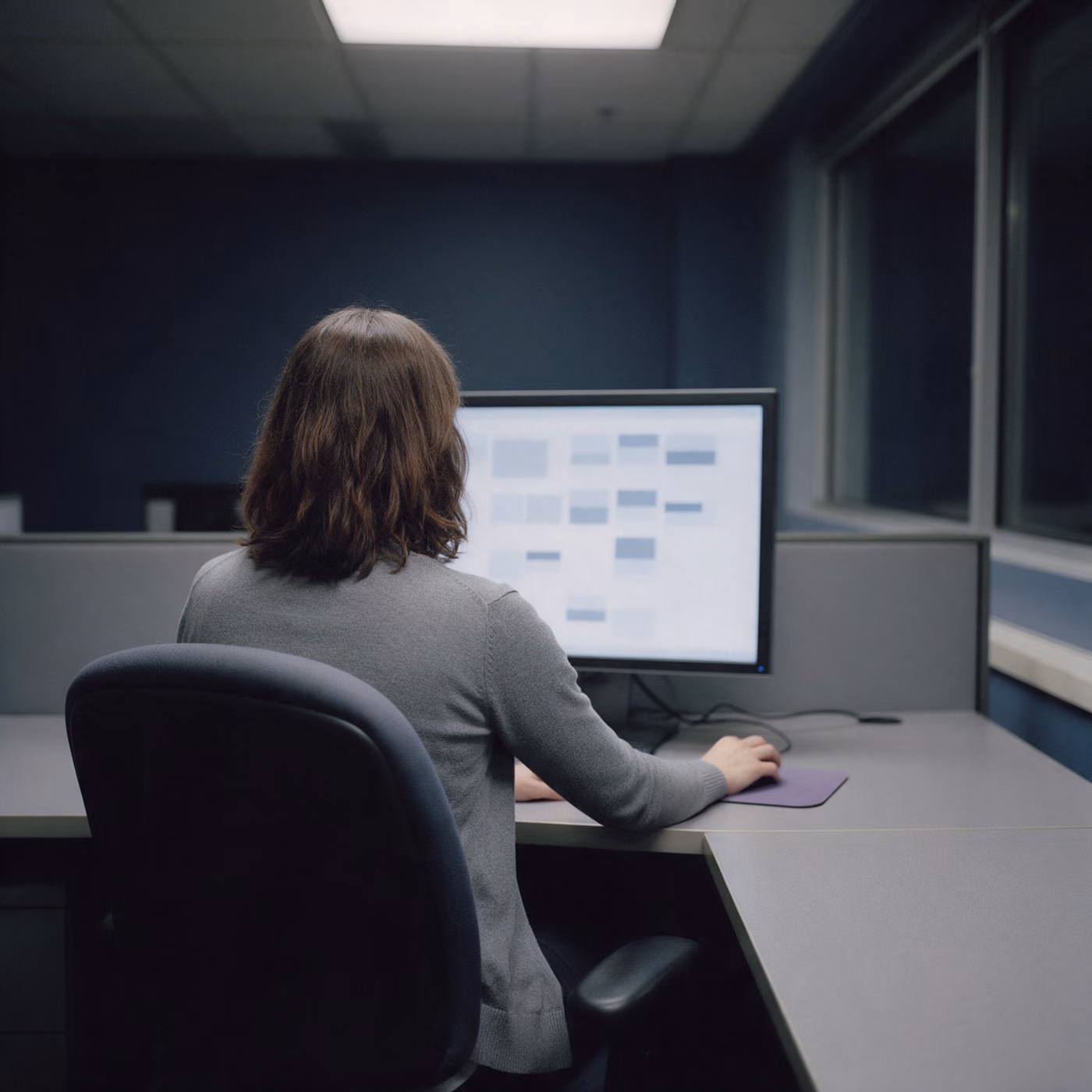 A woman seen from behind at a desk in an office after dark, looking at a large monitor showing a grid of blurred blocks.