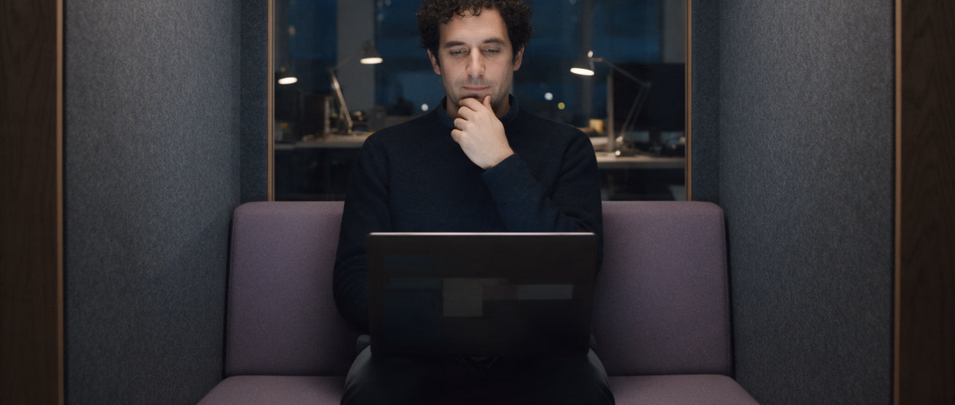 A man sits alone in a felt-lined focus booth after dark, a laptop open on his knees and one hand at his chin.