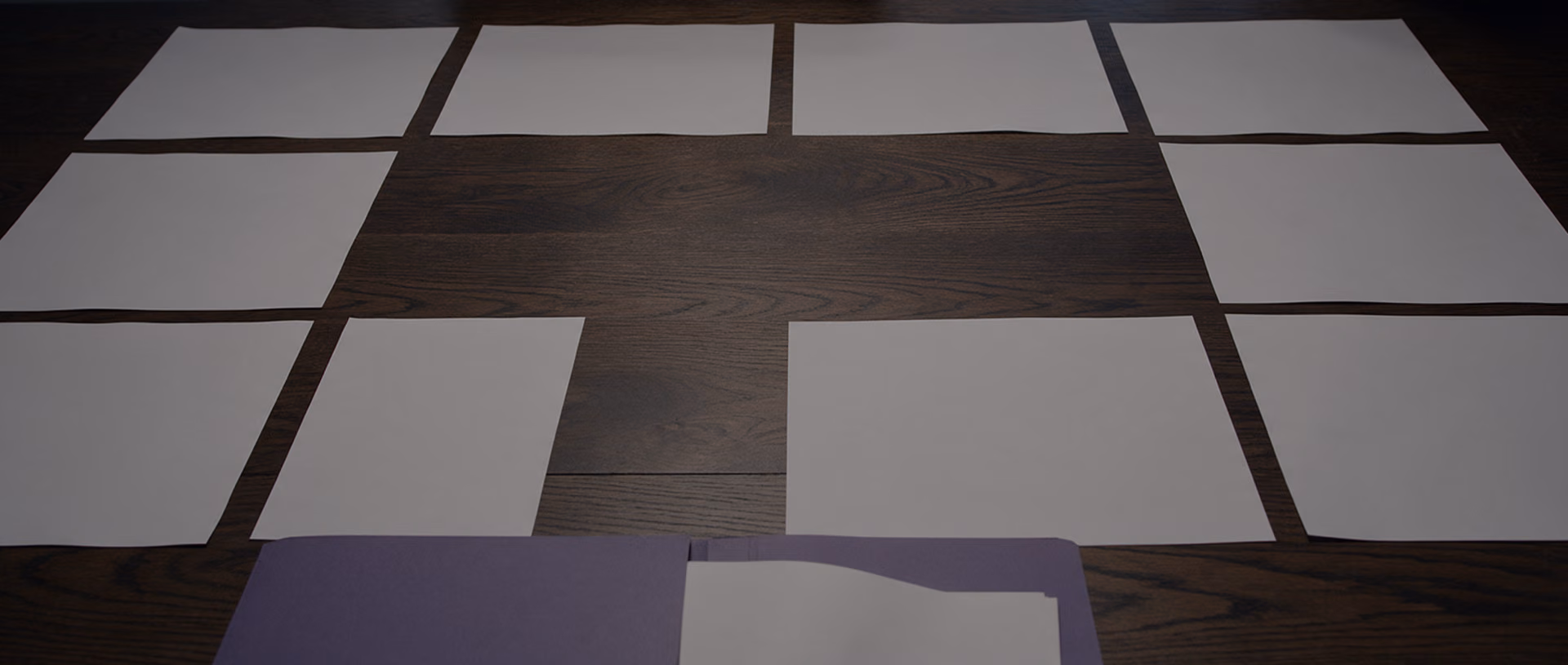 Blank sheets of paper laid out in a grid on a dark table, with several places left empty so that the bare table top shows through.