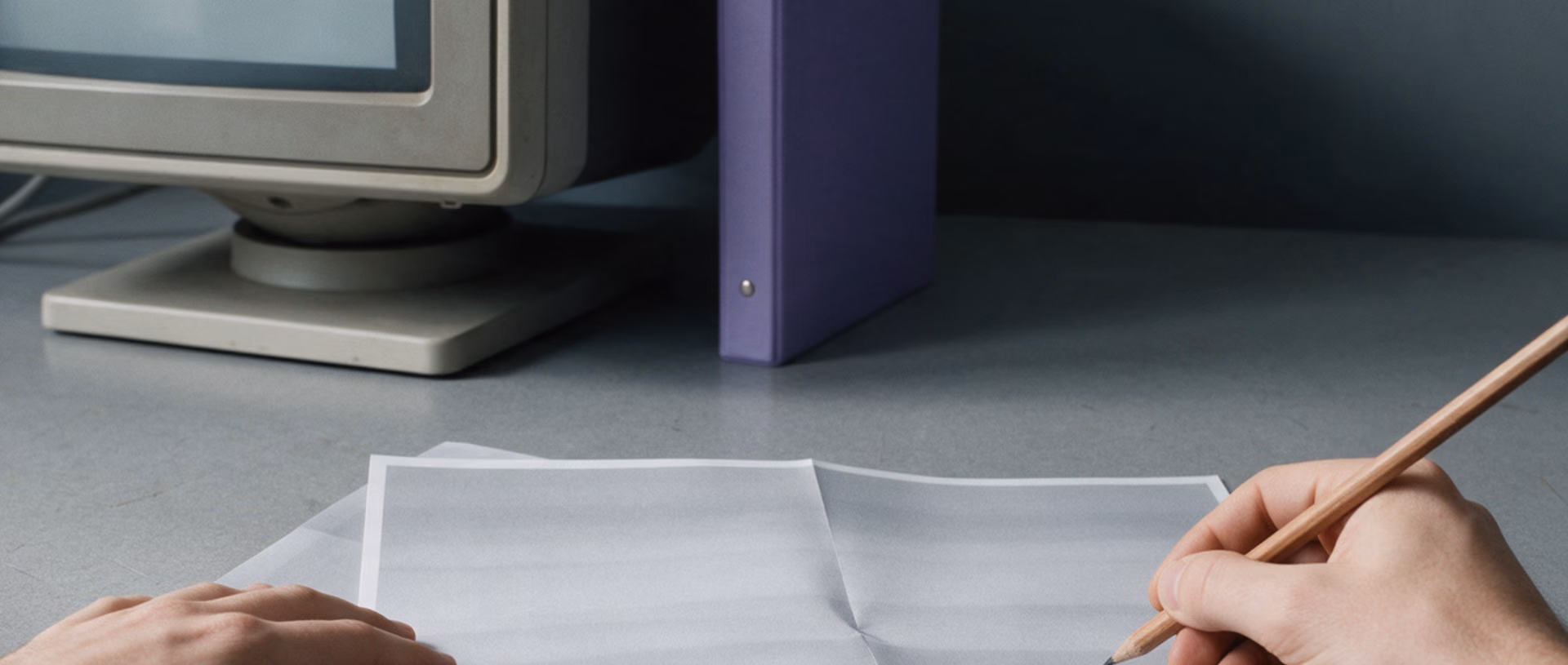 A hand writes with a pencil on a folded printout at a desk beside an old beige office monitor in a bulky plastic bezel.