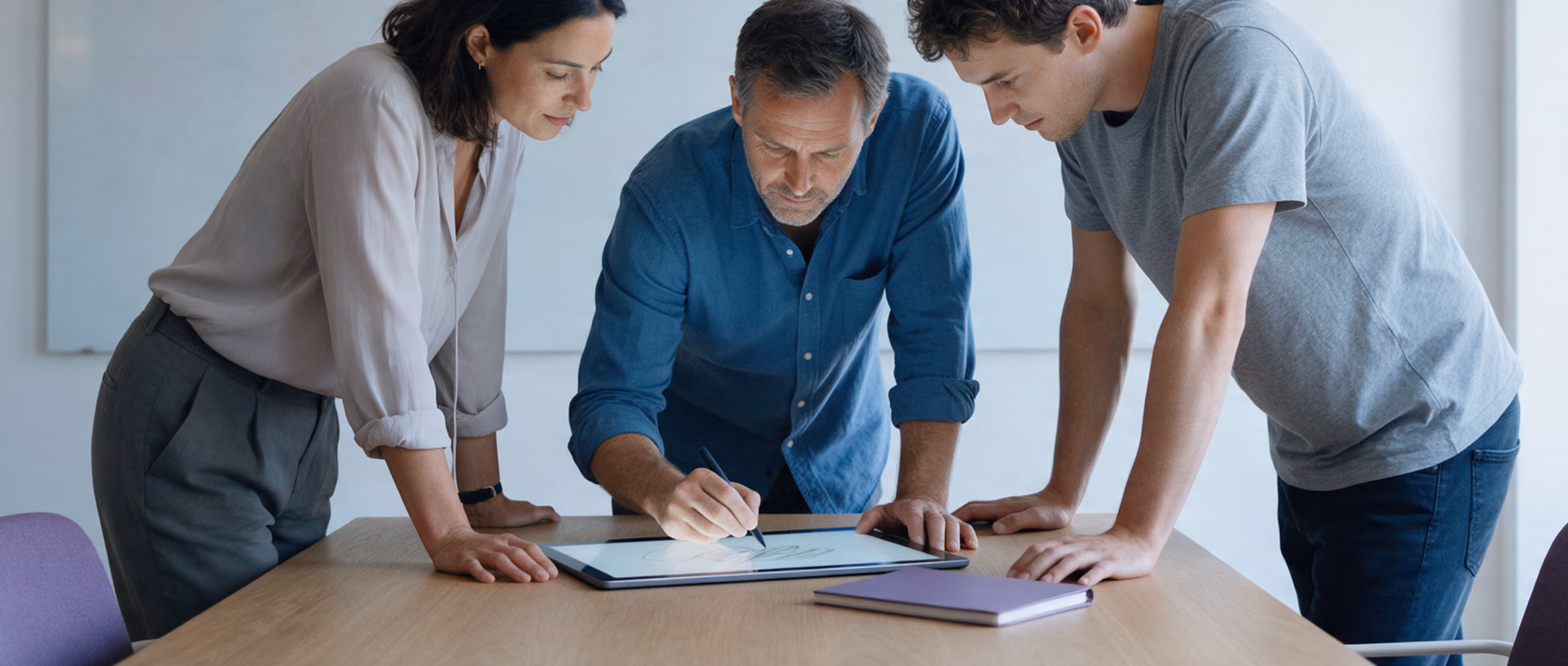 Three colleagues bend over a tall table in a bright project room while one of them sketches on a tablet with a stylus.