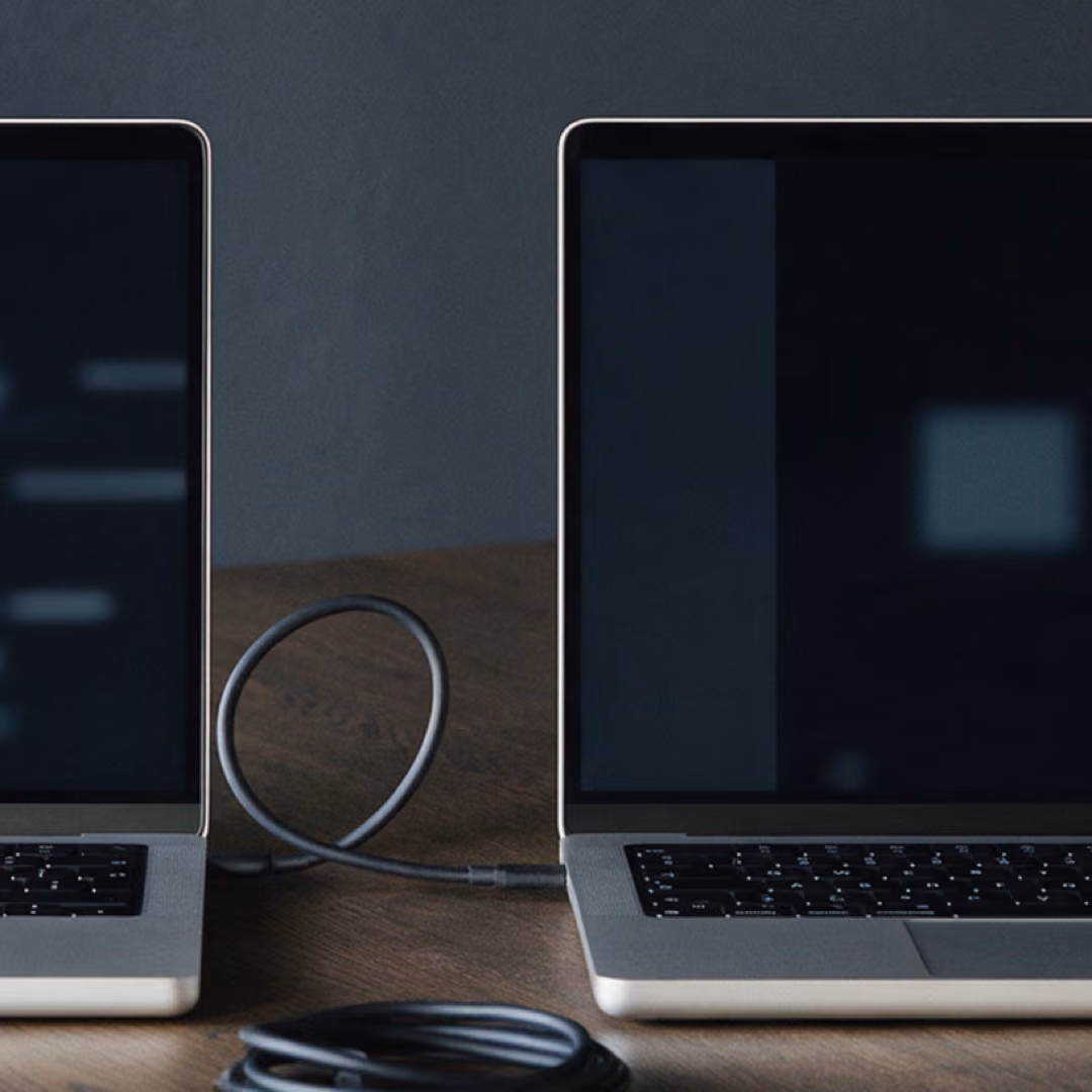 Two laptops side by side on a wooden desk connected by a cable, one screen showing blurred blocks.
