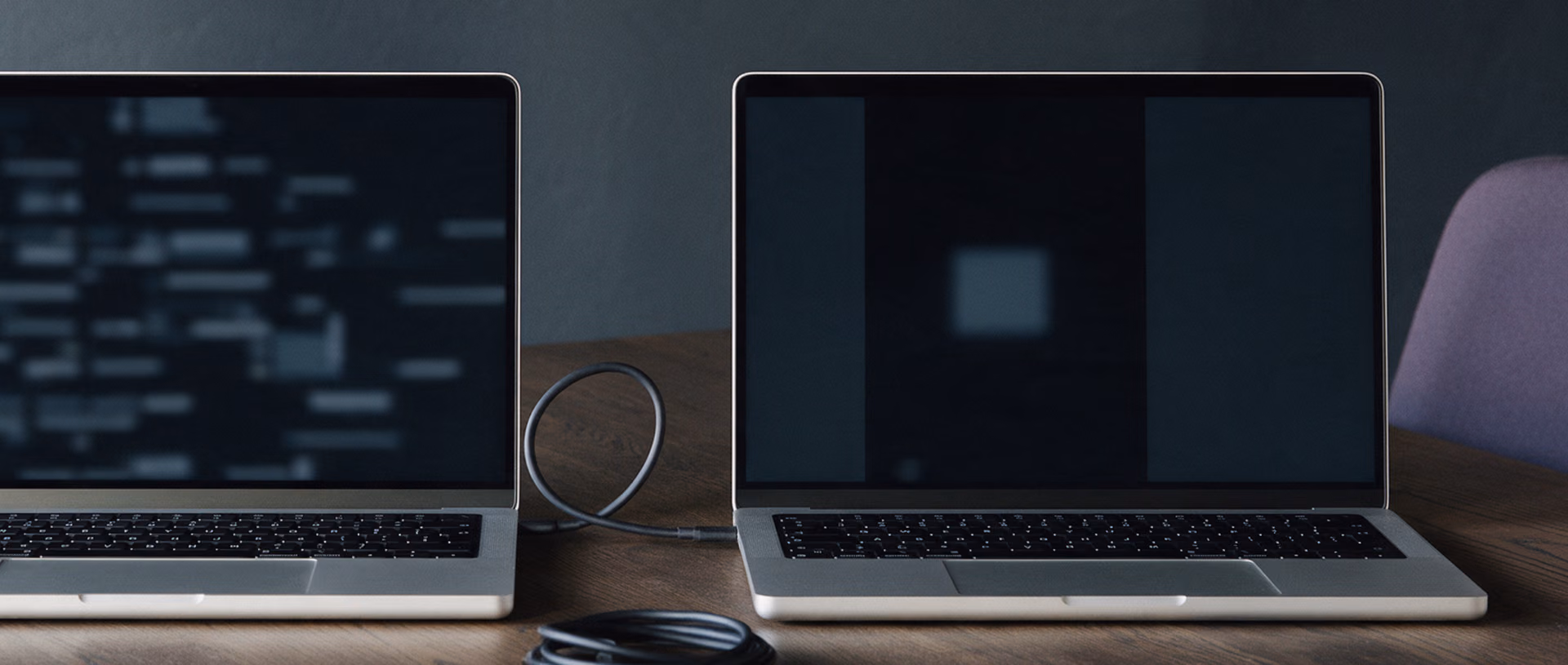 A close view of two laptops side by side on a dark table, joined by a cable, one screen densely filled with blurred blocks and the other nearly empty.