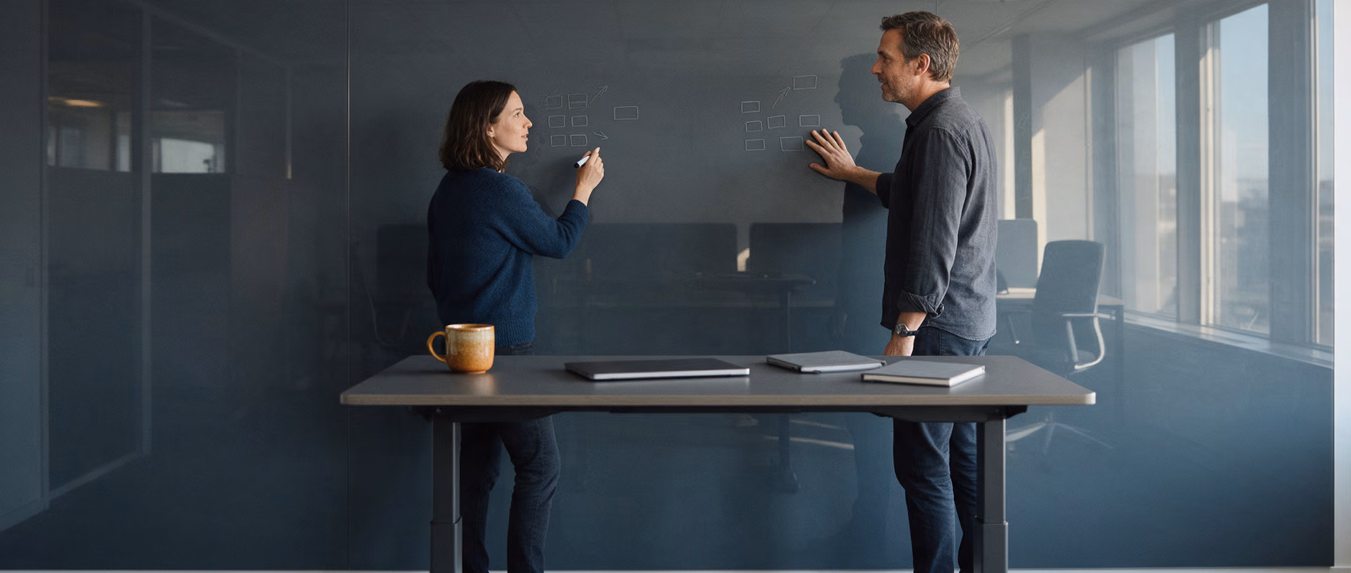 Two colleagues stand at a dark glass writing wall in a project room, each in front of a separate small cluster of sketched boxes, a standing table with a laptop in front of them.