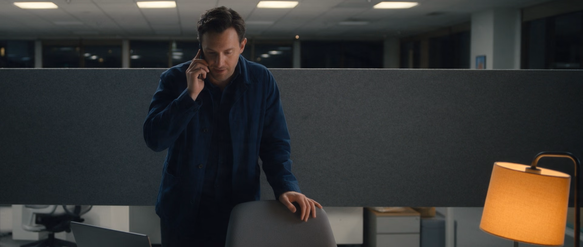 A man in a dark jacket stands at his desk in an office after dark, a phone to his ear, looking down at an open laptop, a felt partition behind him and a lamp at the end of the desk.