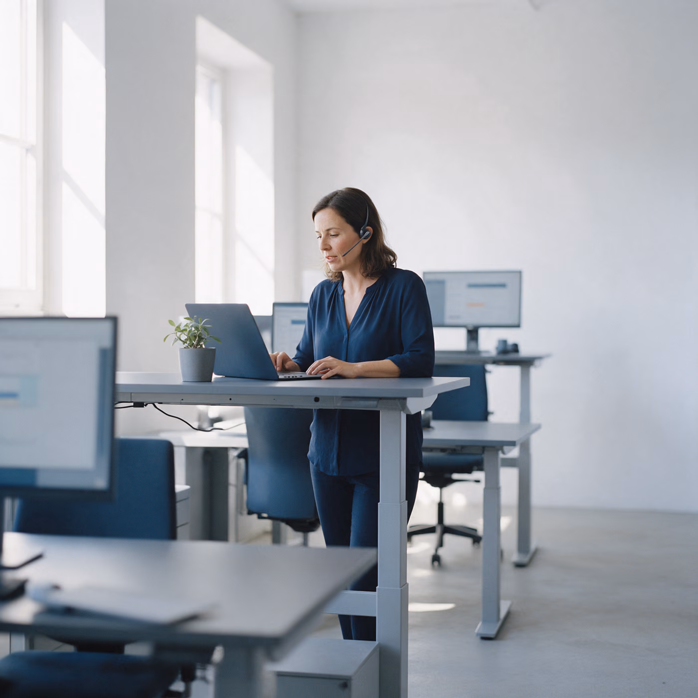 A woman with a headset stands at a height-adjustable desk in a bright office and works on a laptop next to a small potted plant.