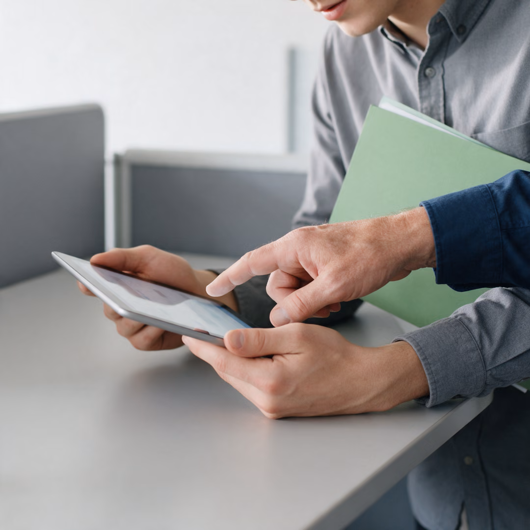 Close view of two people's hands holding a tablet, one finger pointing at the screen, a green folder tucked under one arm.