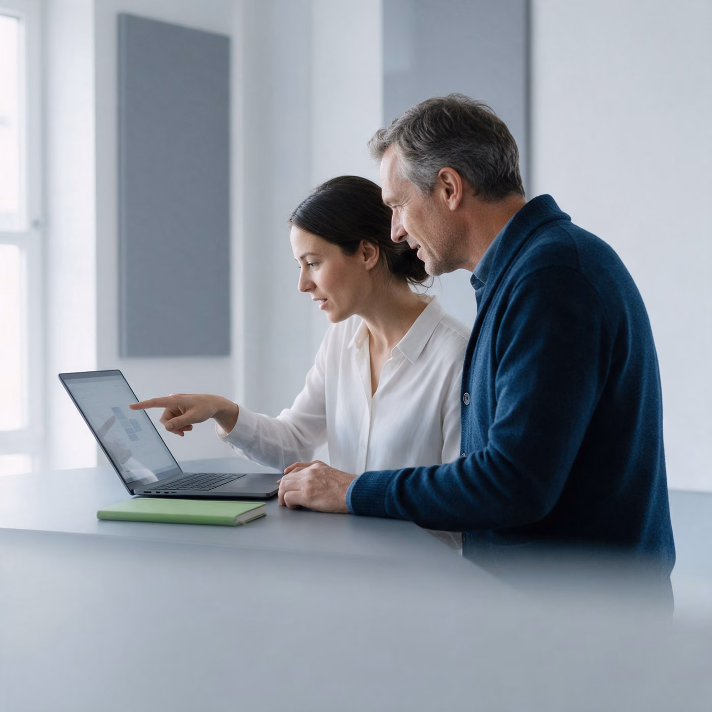 Two colleagues stand at a desk, one pointing at a laptop screen, a green notebook lying beside the laptop.
