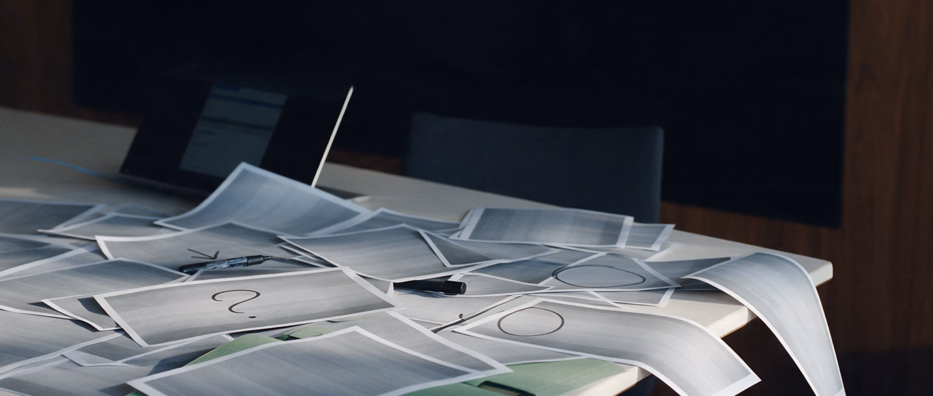 A meeting table strewn with overlapping printed reports in disarray, a few marked by hand with a question mark, a circle or an arrow, a green folder and a half-closed laptop at the table edge.