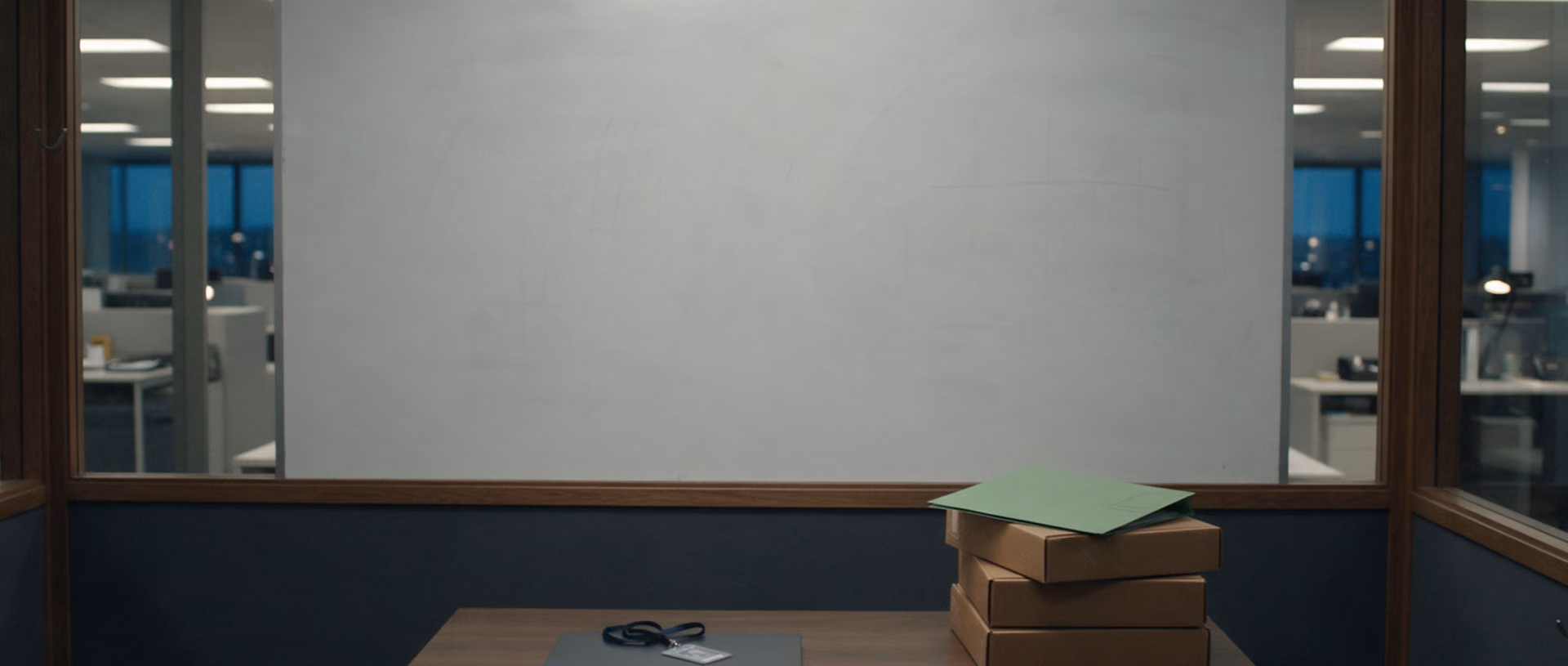 An empty glass-walled side office after hours: a wiped whiteboard above a desk with a closed laptop, a lanyard, a stack of archive boxes and a green folder on top.